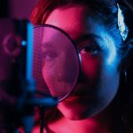 Cantora grava voz diante de um microfone condensador em um estúdio iluminado por luzes coloridas.