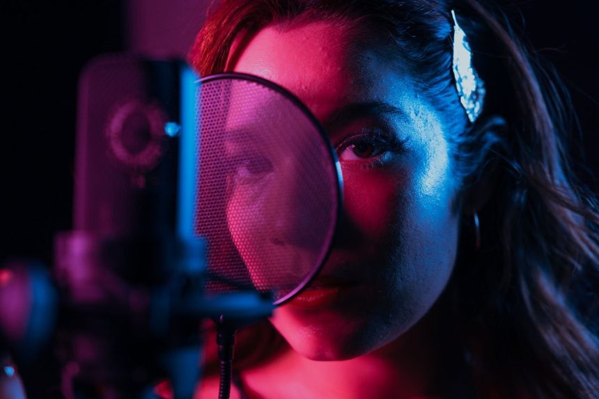 Cantora grava voz diante de um microfone condensador em um estúdio iluminado por luzes coloridas.
