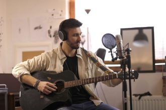 Músico toca guitarra acústica usando fones de ouvido em um estúdio doméstico de gravação.