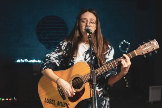 Cantora não identificada se apresenta ao microfone enquanto toca violão em um palco.